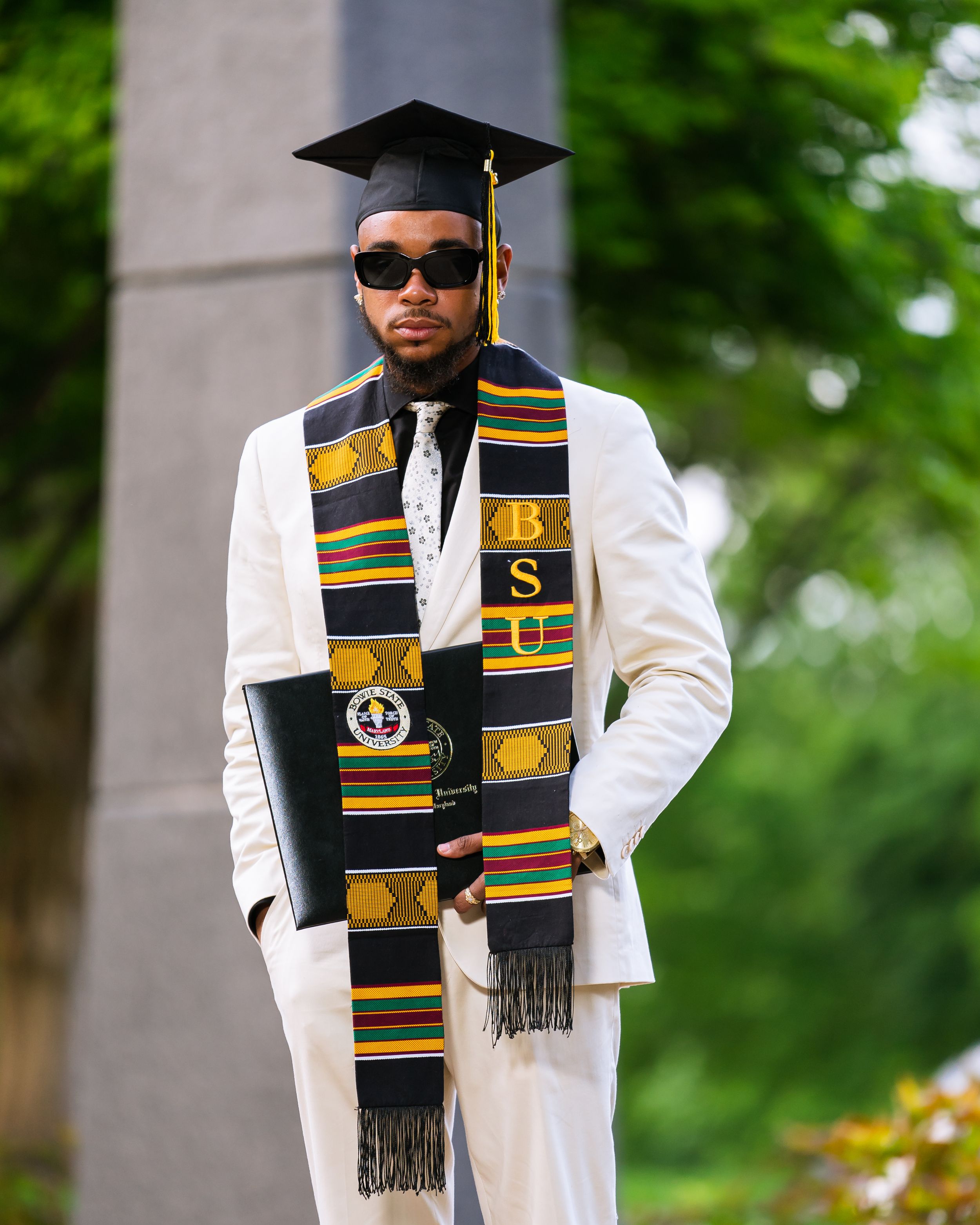 Black male graduate in white suit wearing kente stole and cap holds diploma outdoors.