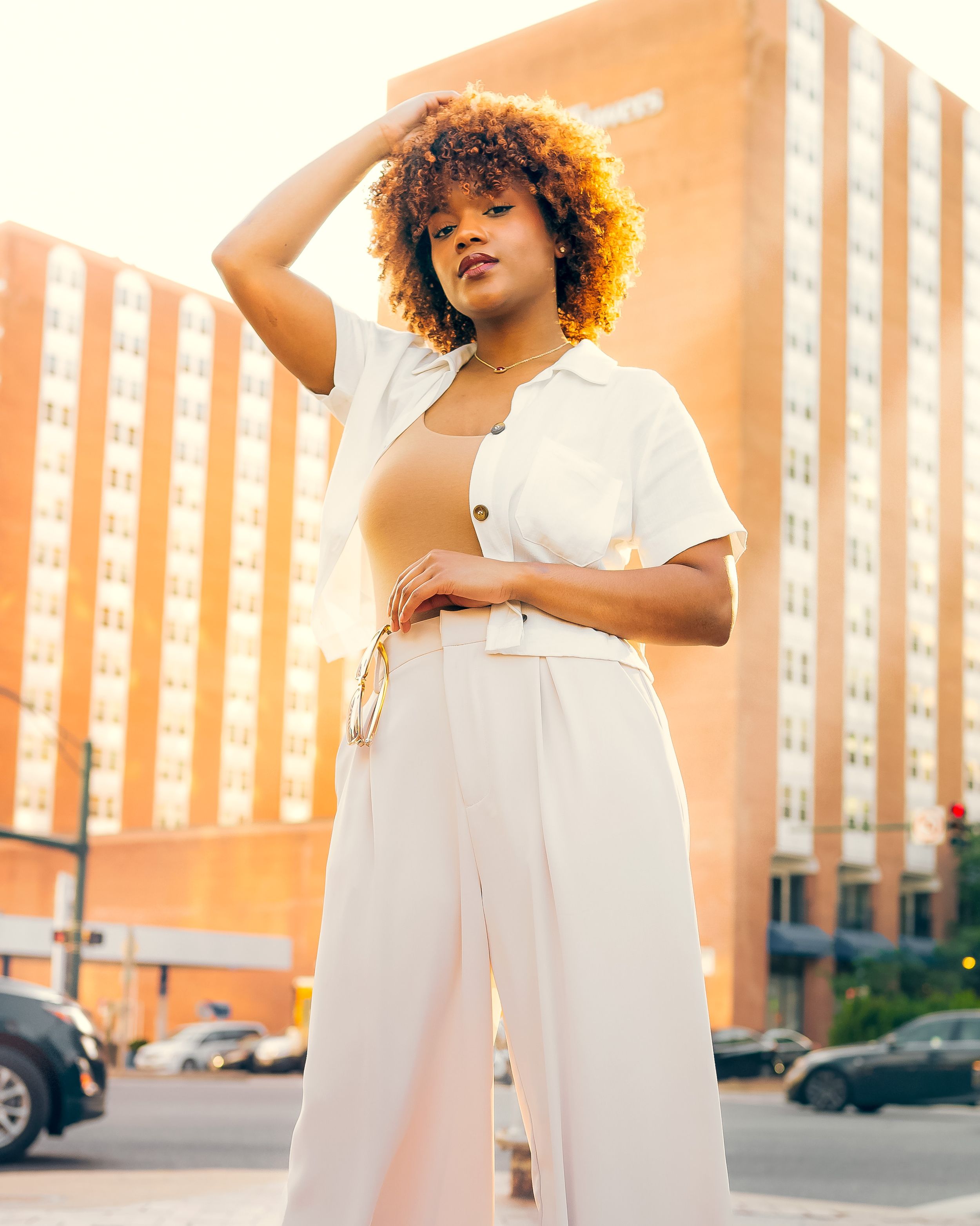 Young woman with curly hair in white wide-leg pants poses on urban street at sunset.