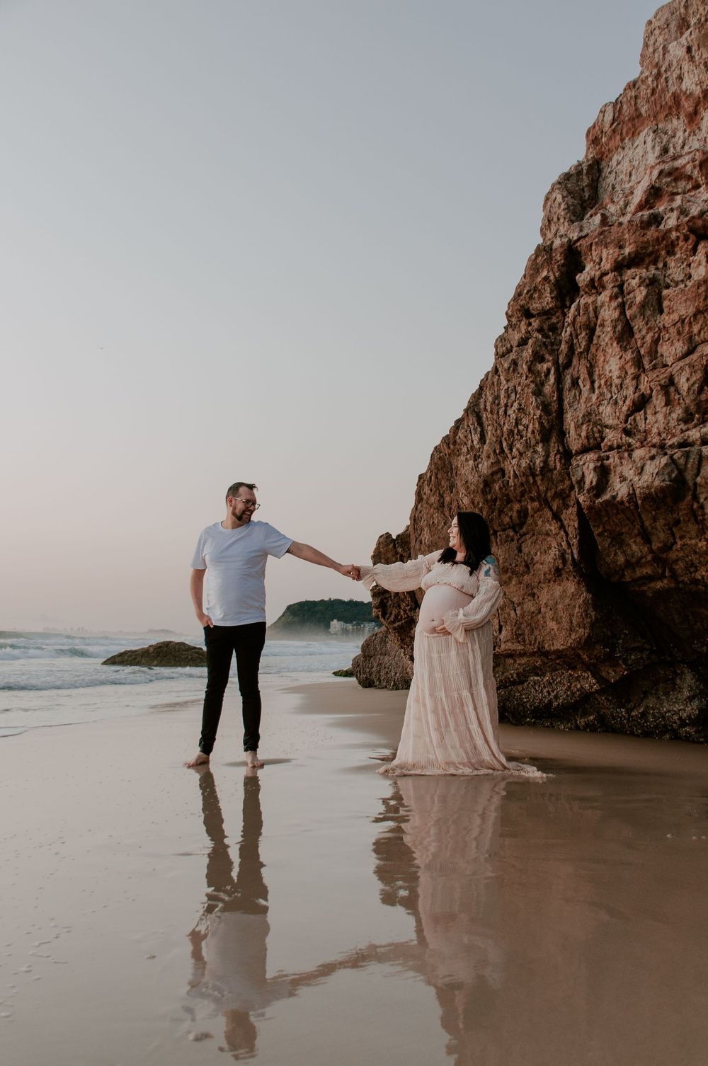 maternity session at the beach. mum and dad to be holding hands and walking towards the camera while waves crash in around them.