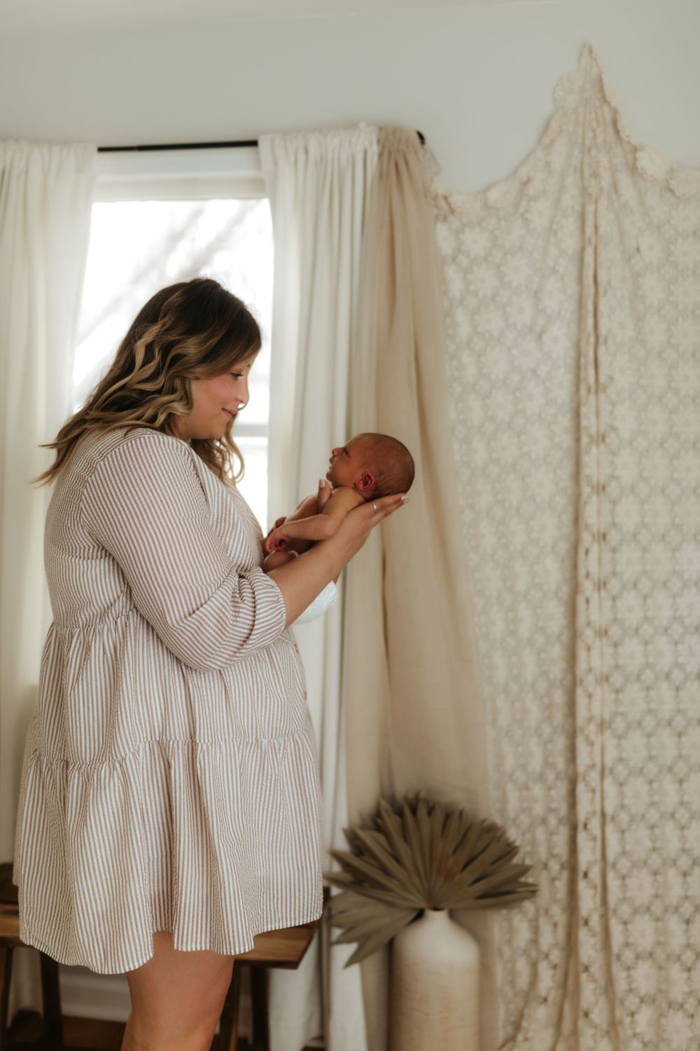 Parent in ribbed dress holding newborn baby by window with decorative screen and palm fan.