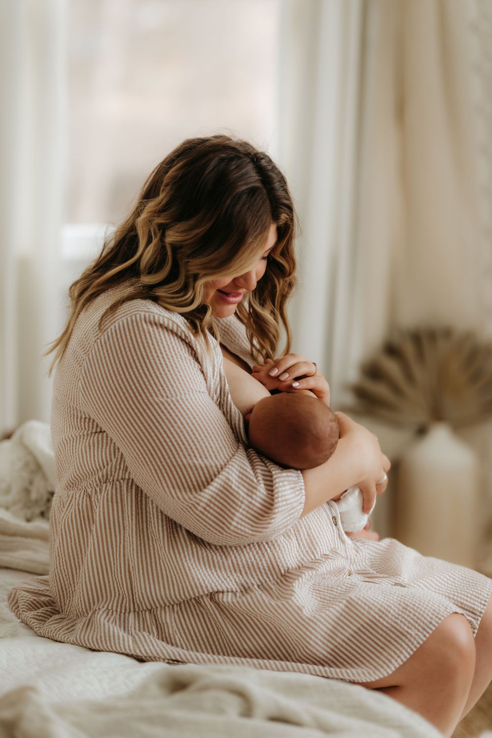 Parent in ribbed sweater dress holding sleeping infant on white bedding with neutral decor.