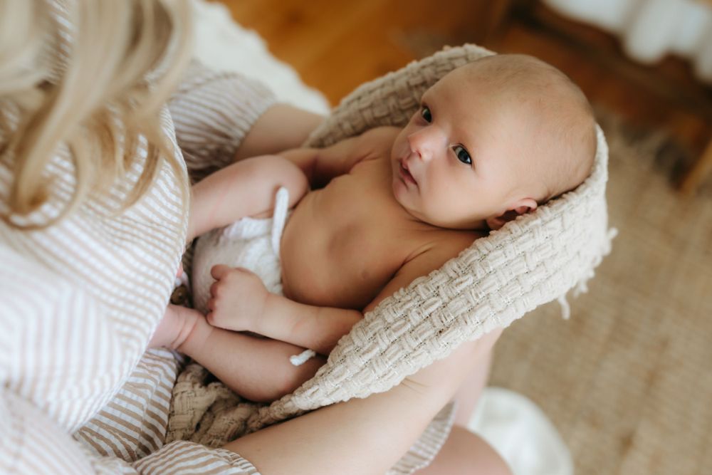 Newborn baby wrapped in knit blanket being held against cream sweater in natural light.