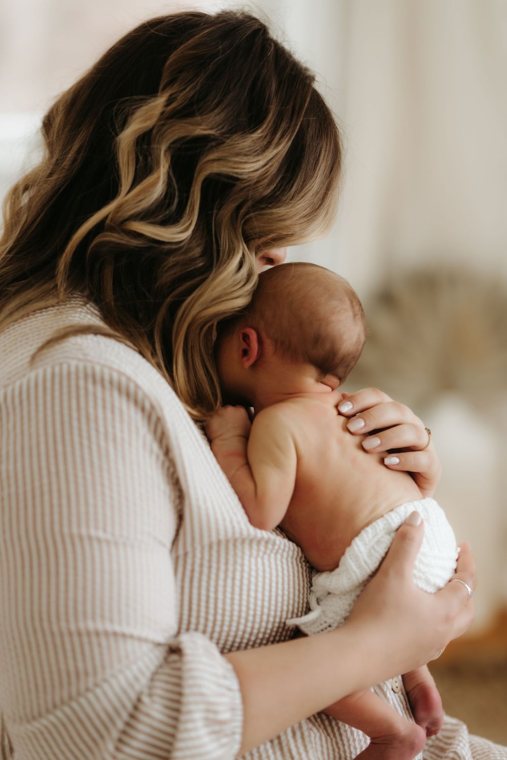 Parent in knit sweater holding sleeping newborn baby in white diaper against light backdrop.
