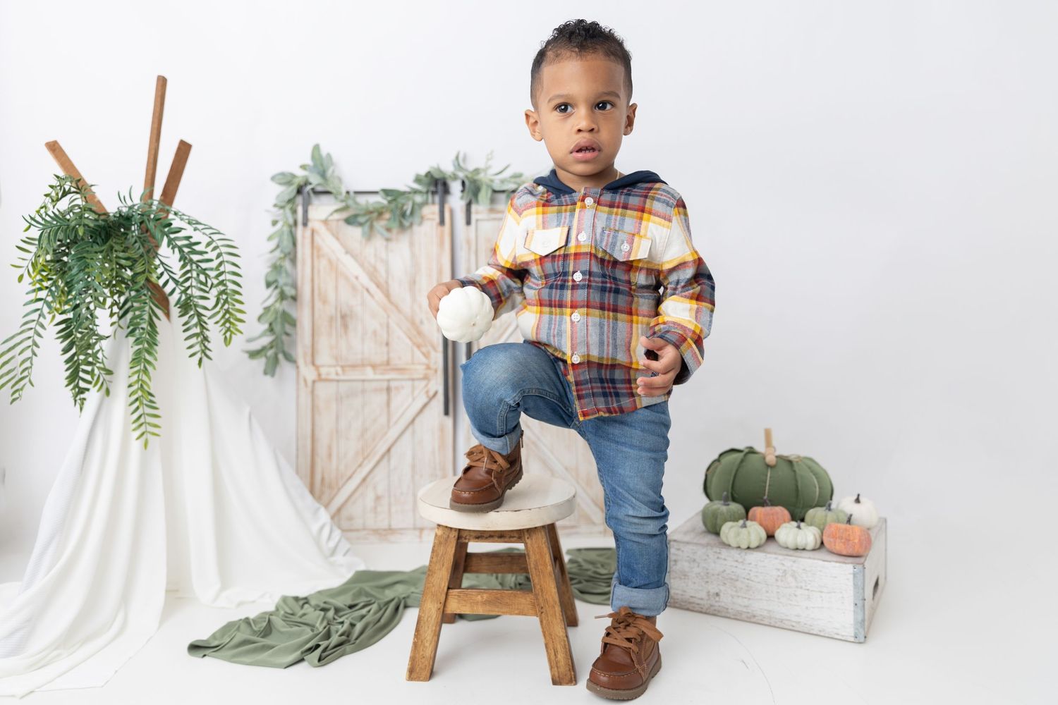 Fall photo setup with wooden stool, rustic door, pumpkins and hanging greenery in natural light.