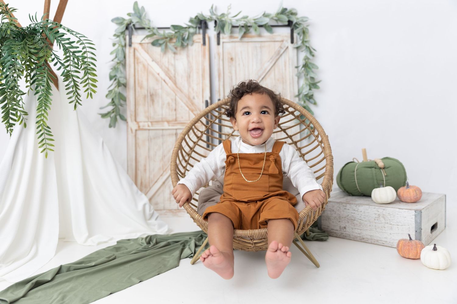 Rustic photo setup with rattan chair, barn door backdrop, green pumpkins, and hanging ferns in natural tones.