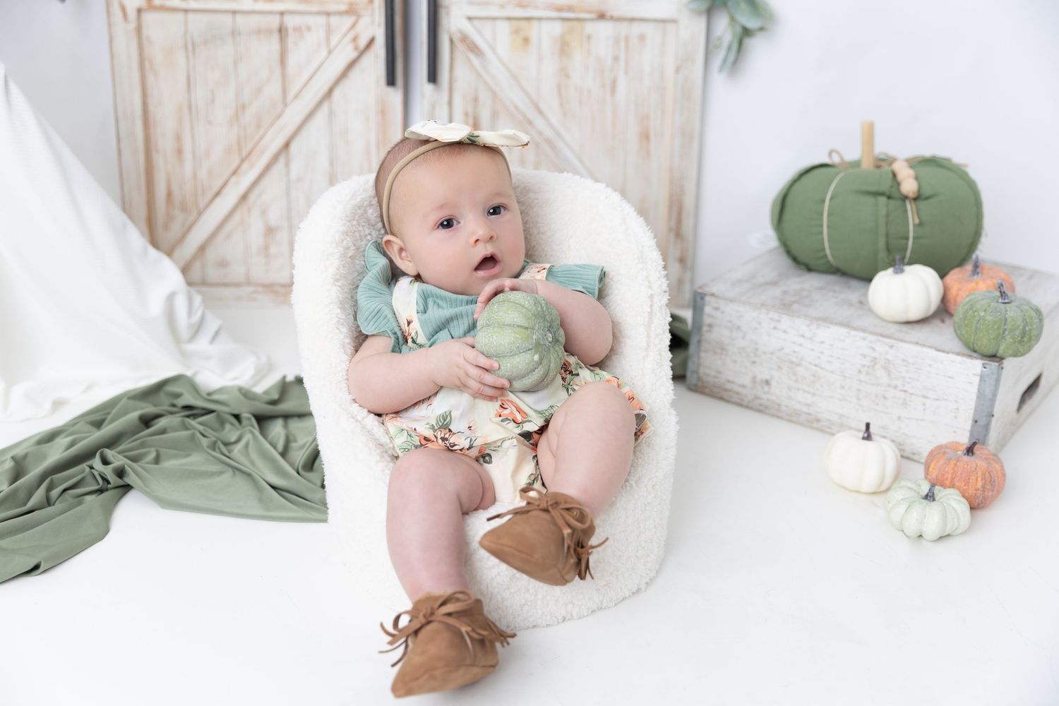 Minimalist photo setup with white cushioned chair, rustic backdrop and green pumpkin decorations.