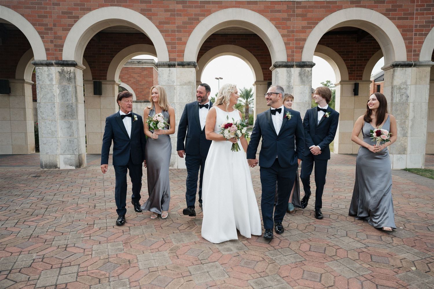 Wedding party walks together under a series of brick arches on a paved courtyard.