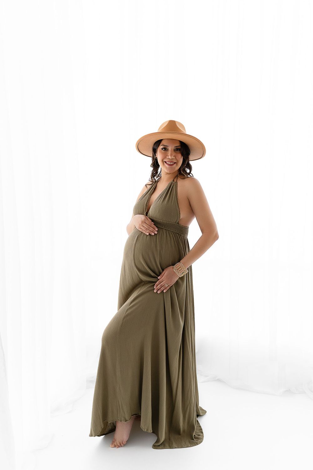 Maternity portrait in olive maxi dress and tan hat against white backdrop.