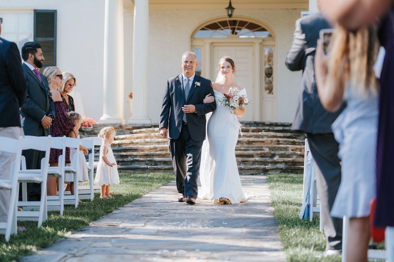 Wedding ceremony processional on stone pathway with white chairs and guests.