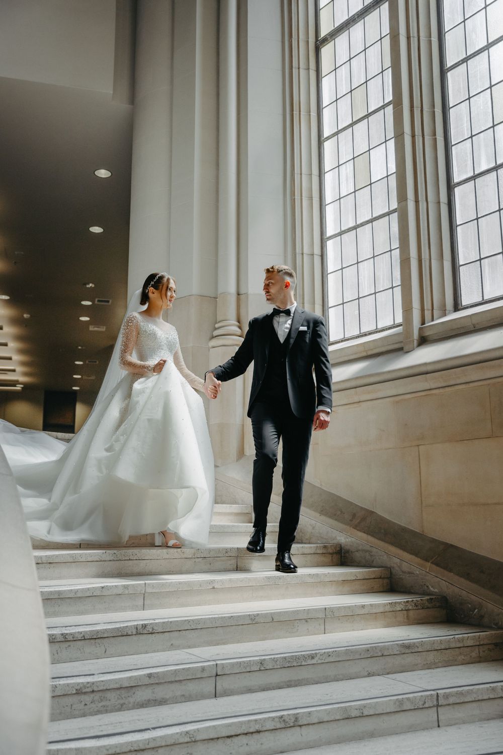 Wedding couple descending marble stairs near tall church windows in an elegant interior.
