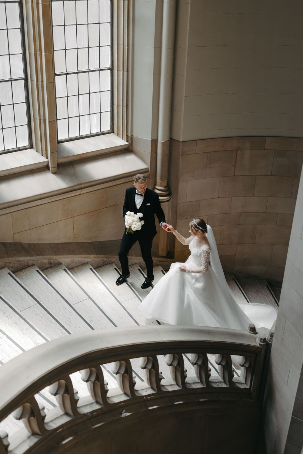 Wedding couple poses on grand staircase with architectural details and window light.