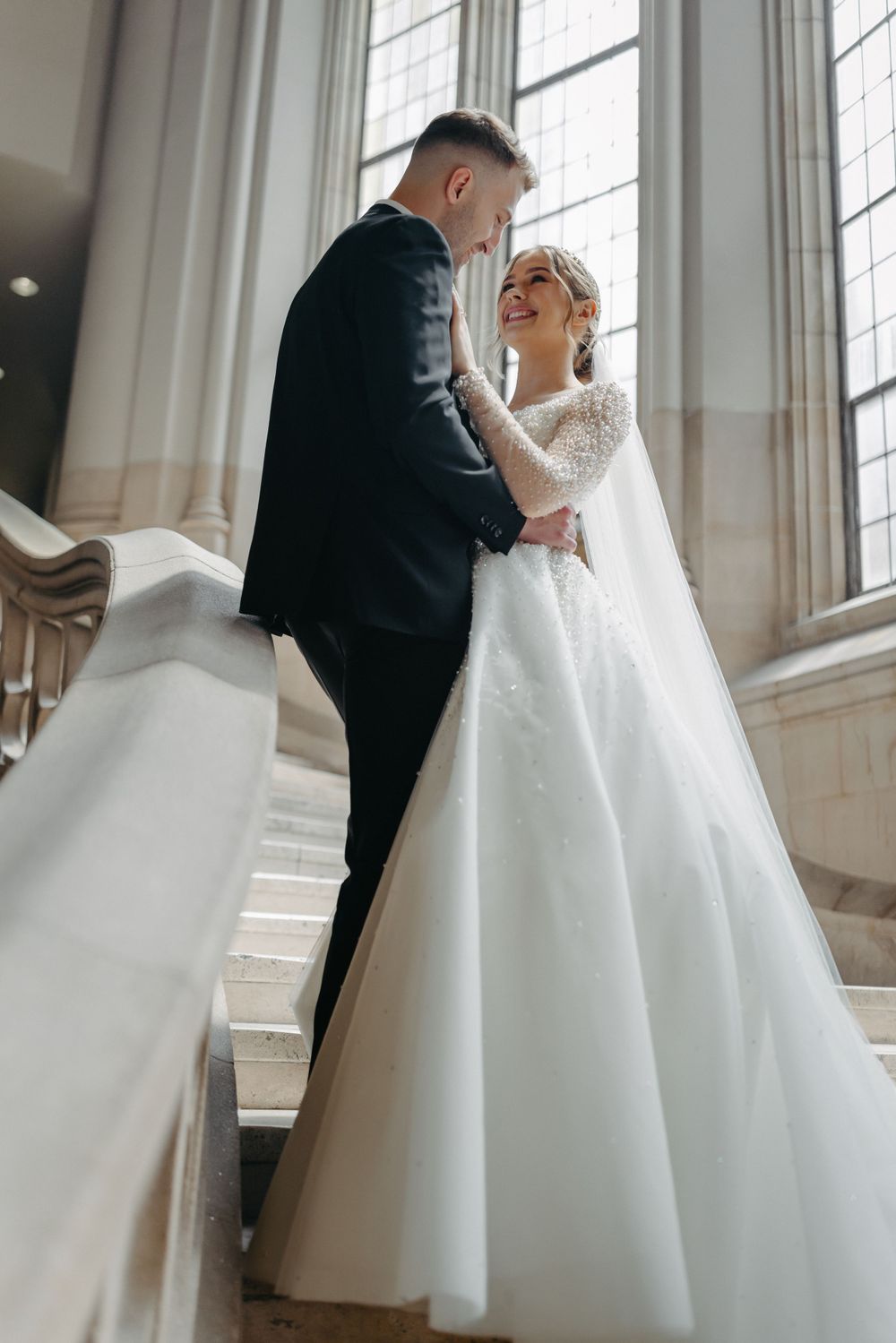 Romantic staircase wedding portrait with flowing white gown and tall windows.