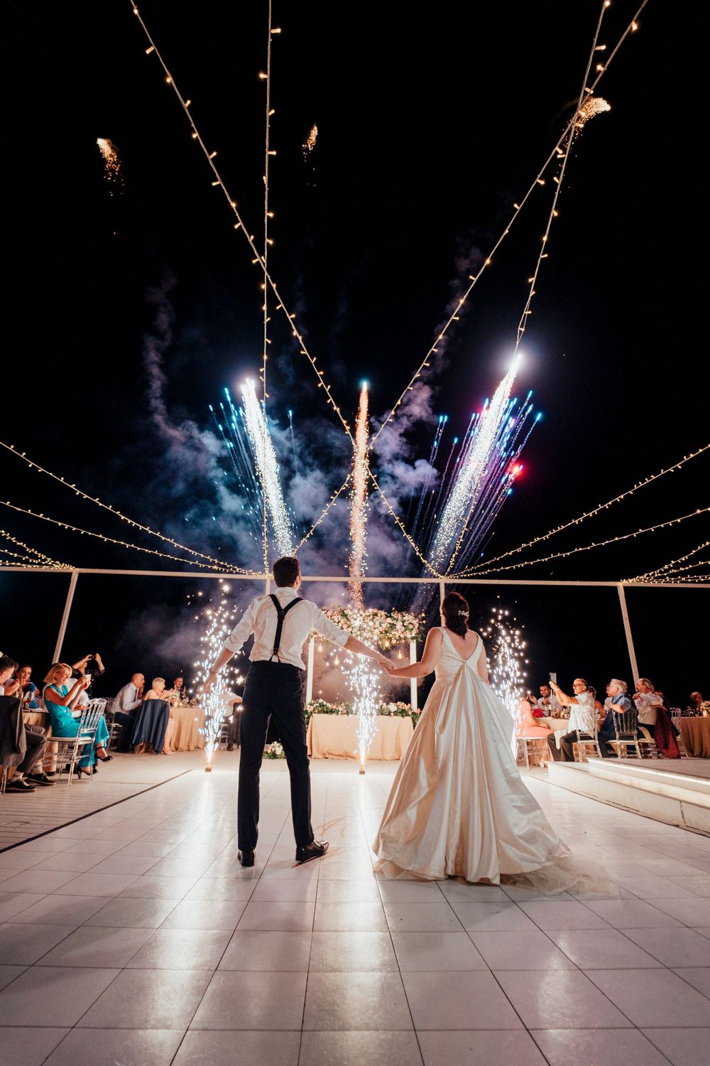 Colorful fireworks light up the night sky during a wedding dance on an outdoor patio.