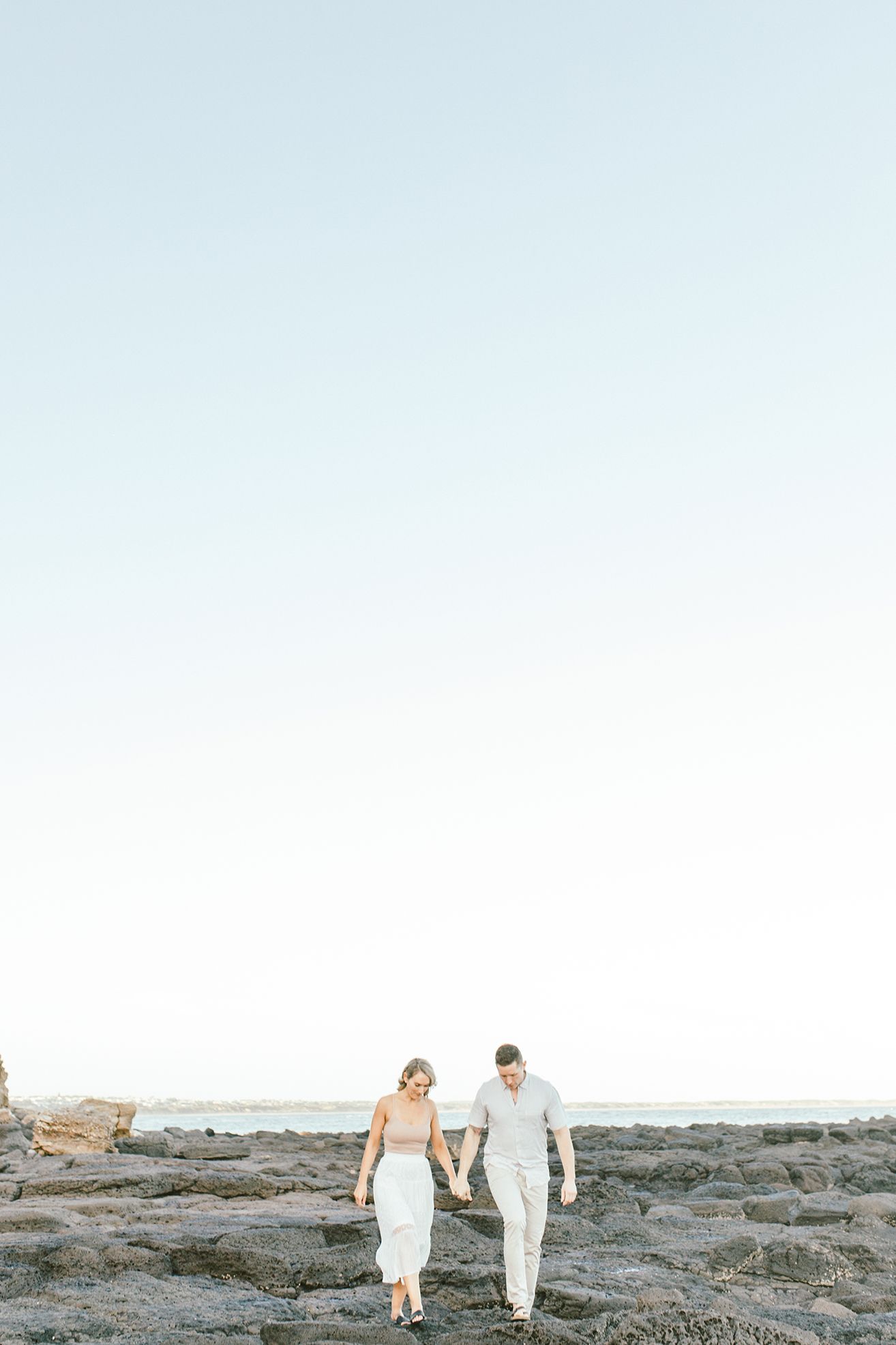 Couple walking together on rocky coastal terrain during sunset.