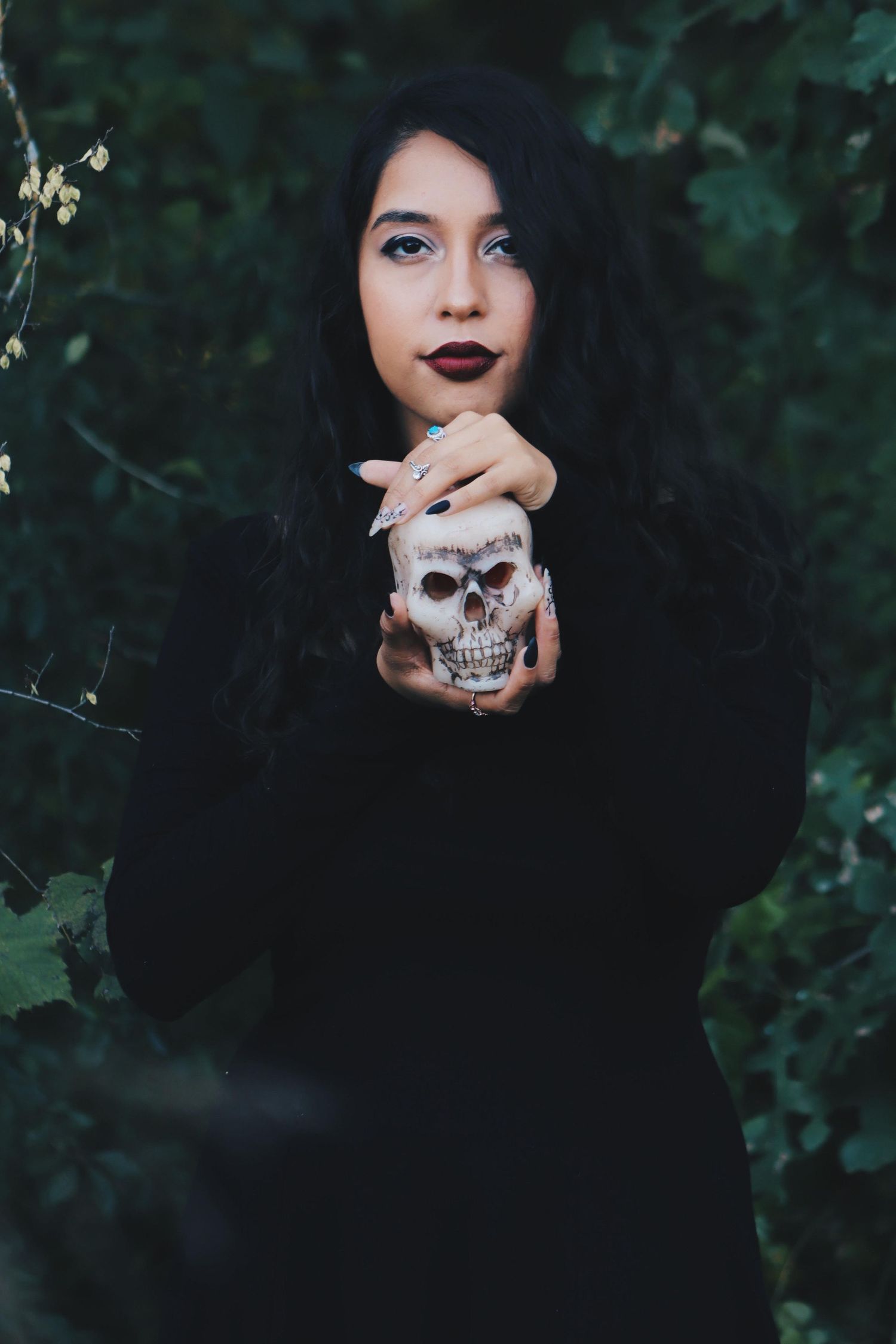 Young woman in black outfit holding decorative skull prop surrounded by green leaves in moody Halloween-themed portrait.