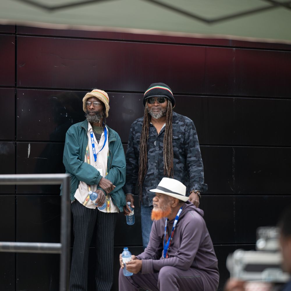 A person in a light-colored hat sits on a bench while others stand nearby during what appears to be an outdoor event.