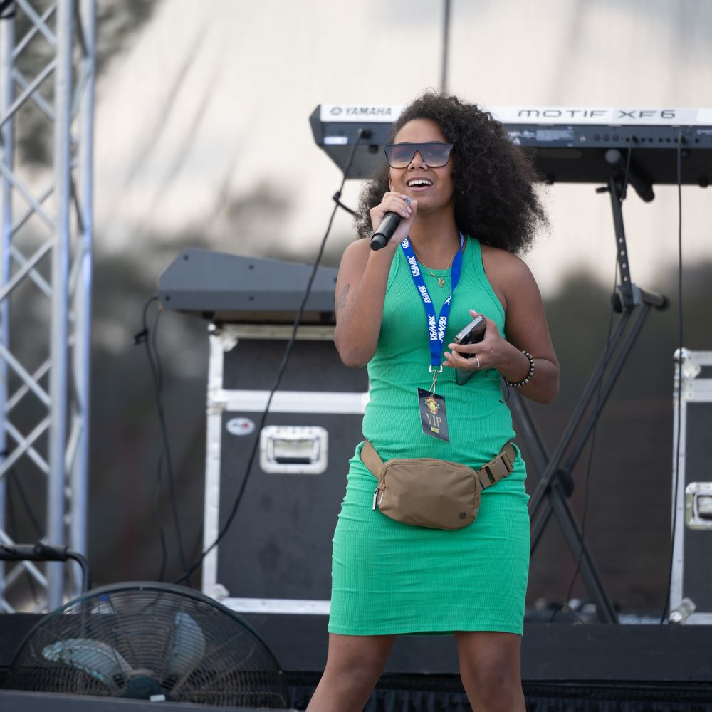 Performer in a bright green tank top sings energetically on an outdoor stage with sound equipment visible.