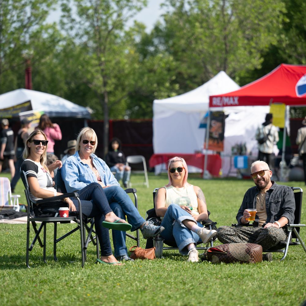 People relax on lawn chairs at an outdoor festival with red tents visible in the background on a sunny day.