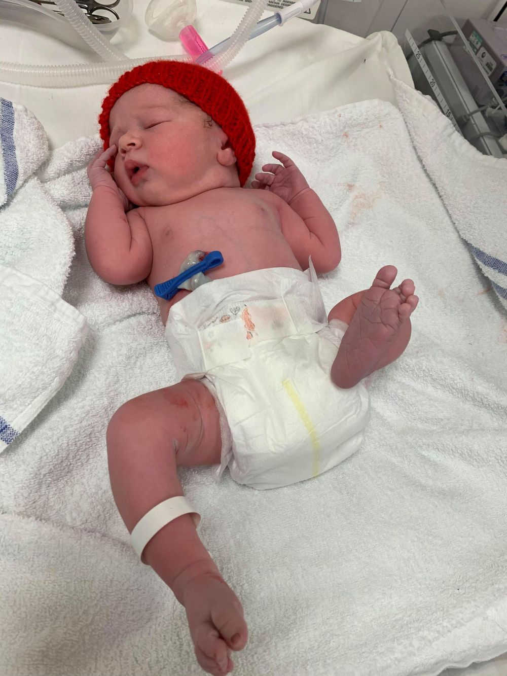 Newborn wearing red knit hat resting on white hospital blanket.
