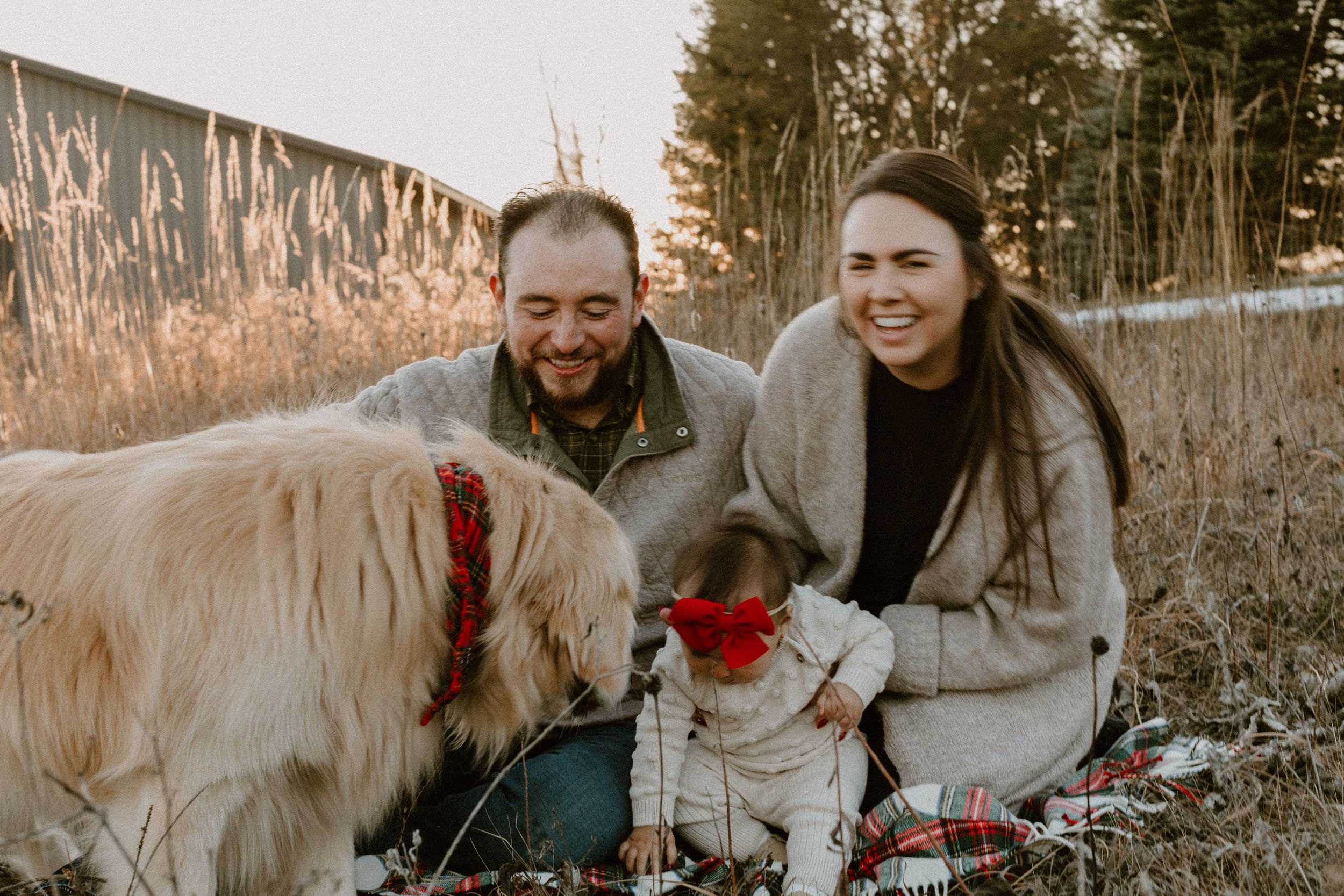 Family and their golden retriever dog wearing a red bow outdoors in a natural winter setting.