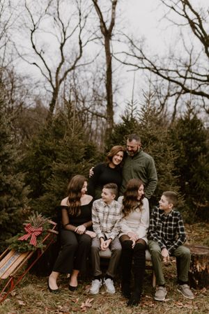 Family of six posed together on wooden bench surrounded by evergreen trees.