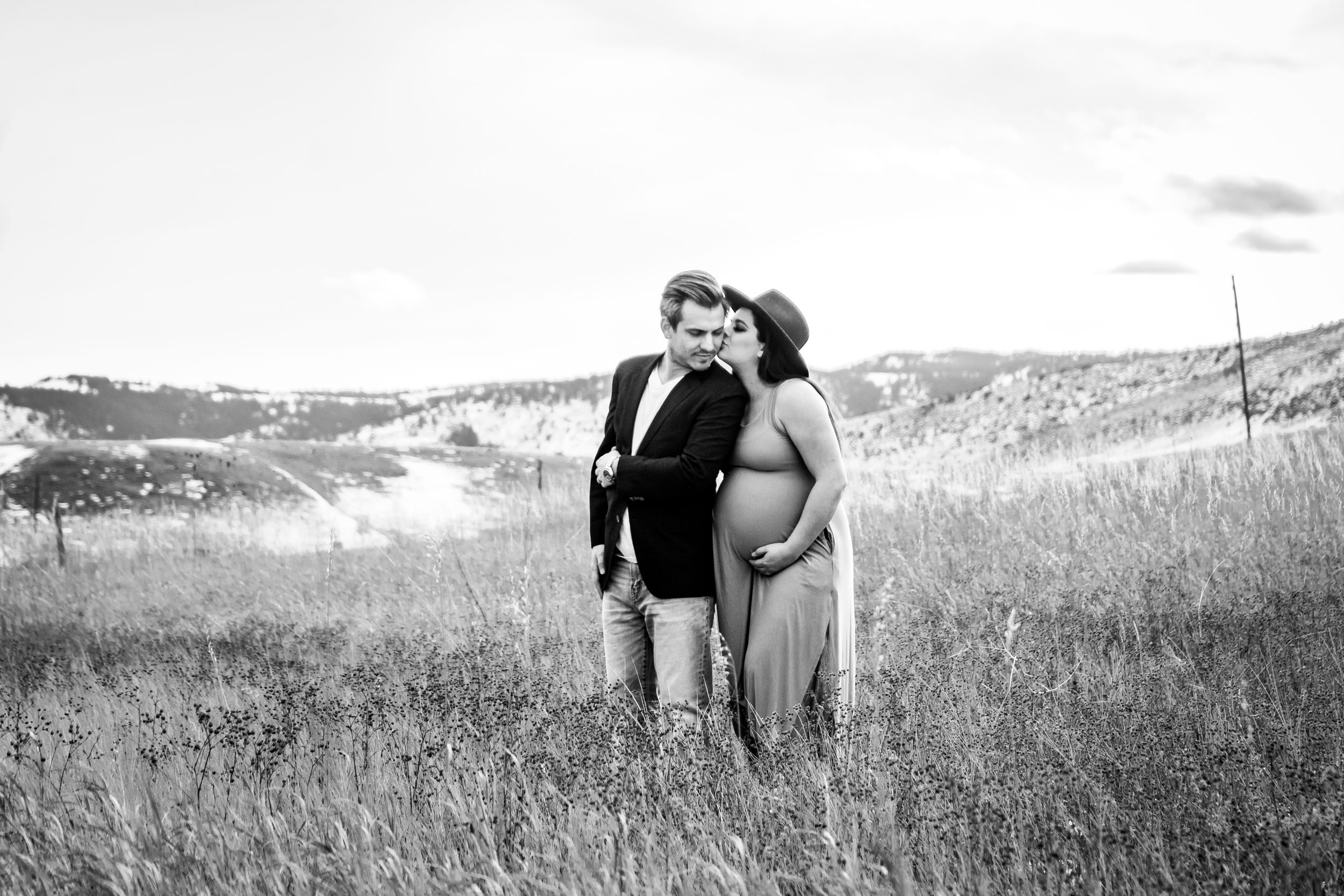 Black and white image of a father and pregnant mother standing together in a grassy field with rolling hills in the distance.