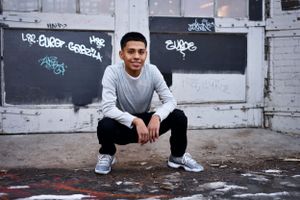Urban high school senior portrait against graffiti wall wearing grey sweater, black pants, and sneakers.