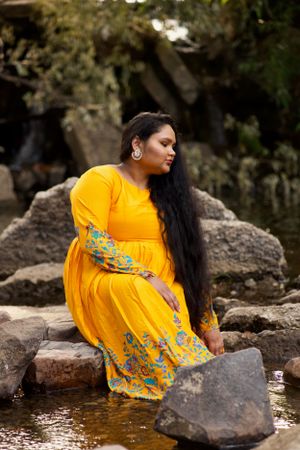 Woman in yellow sari with floral embroidery photographed near rocks and water in an outdoor setting.