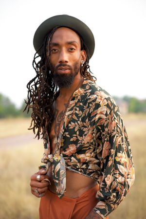 Outdoor fashion portrait of a man in floral print shirt and rust colored pants with long braids and wide brim hat.