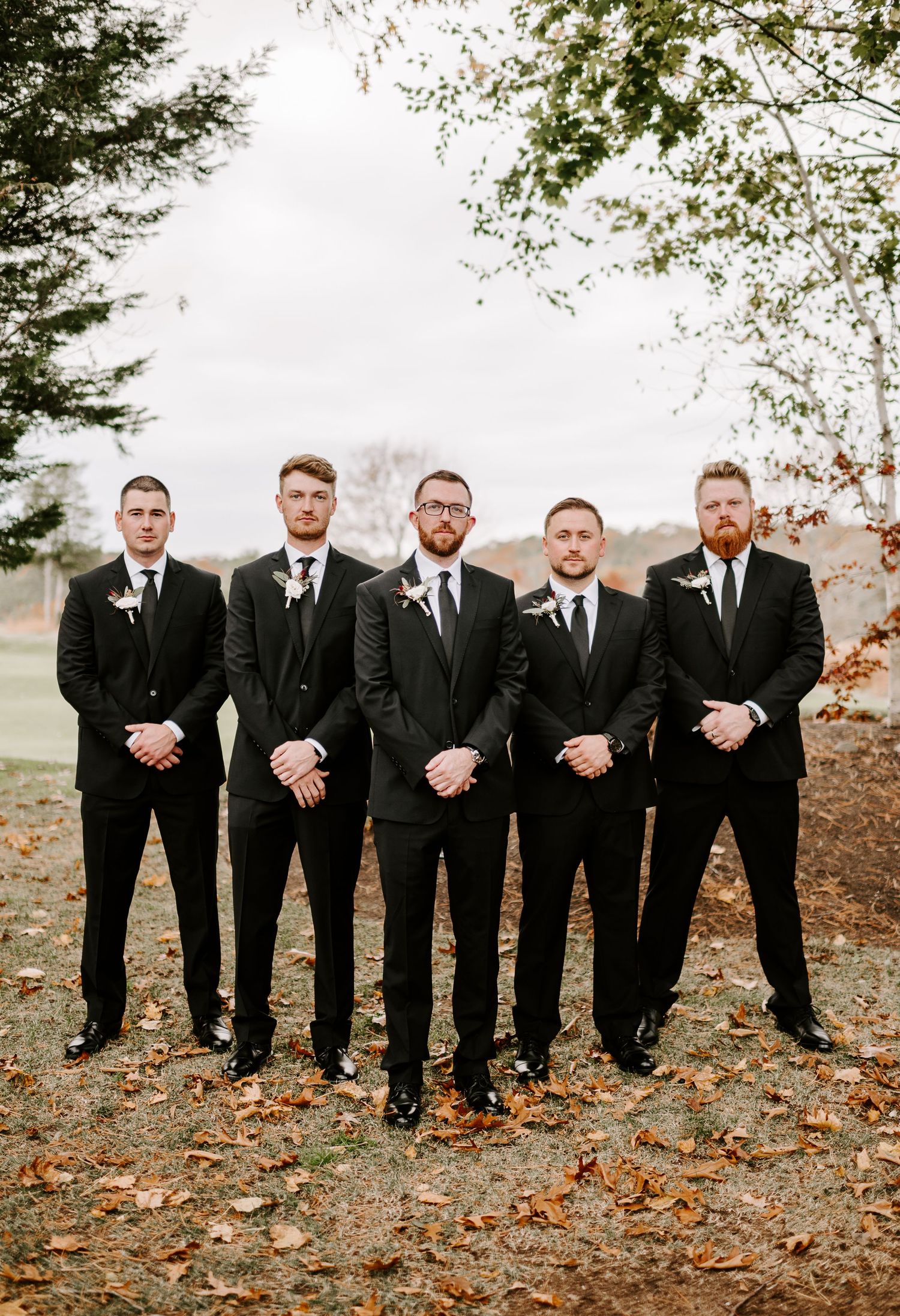 Groomsmen in black suits pose formally against autumn landscape with fallen leaves.
