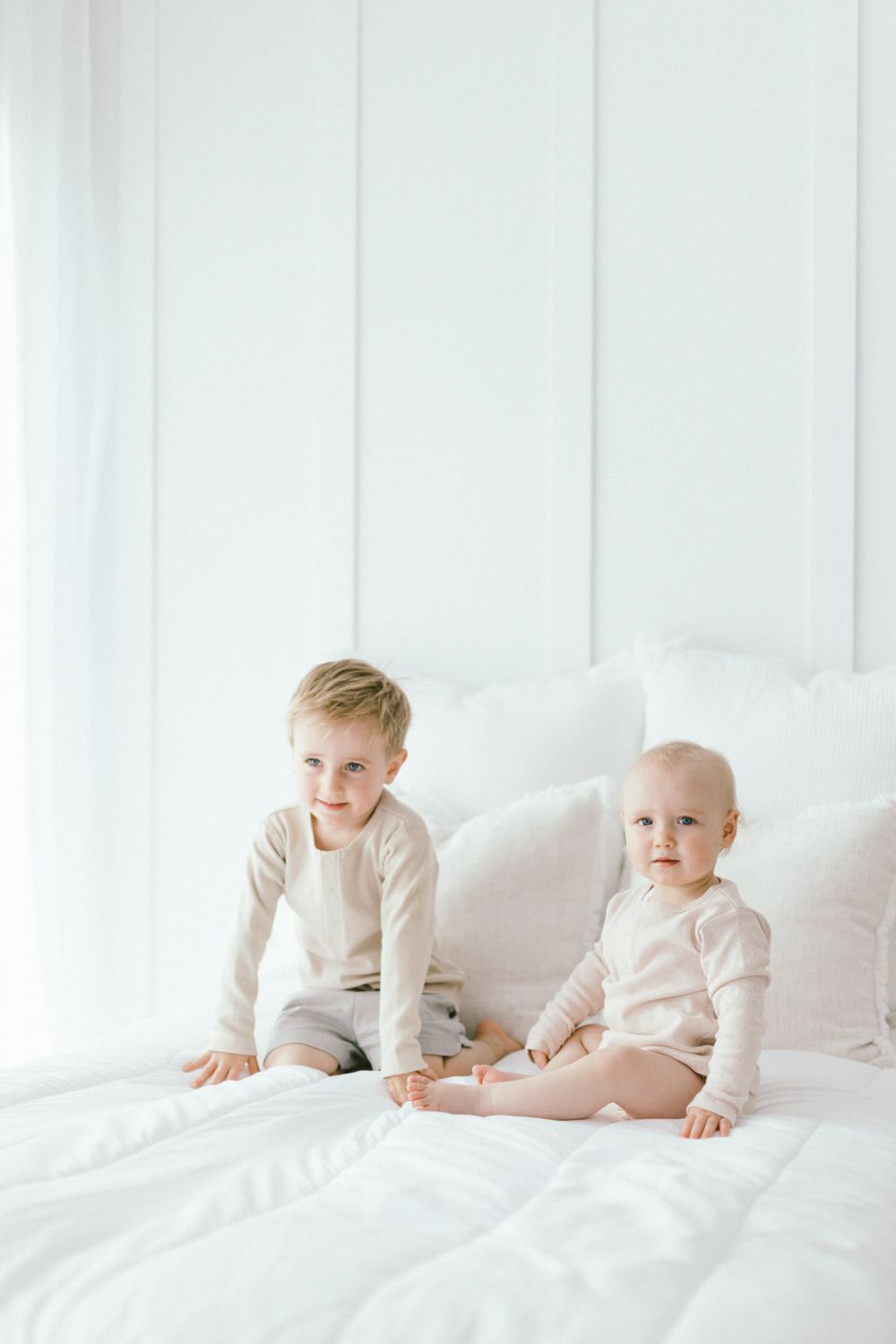 Two babies in light-colored outfits sitting on white bedding.