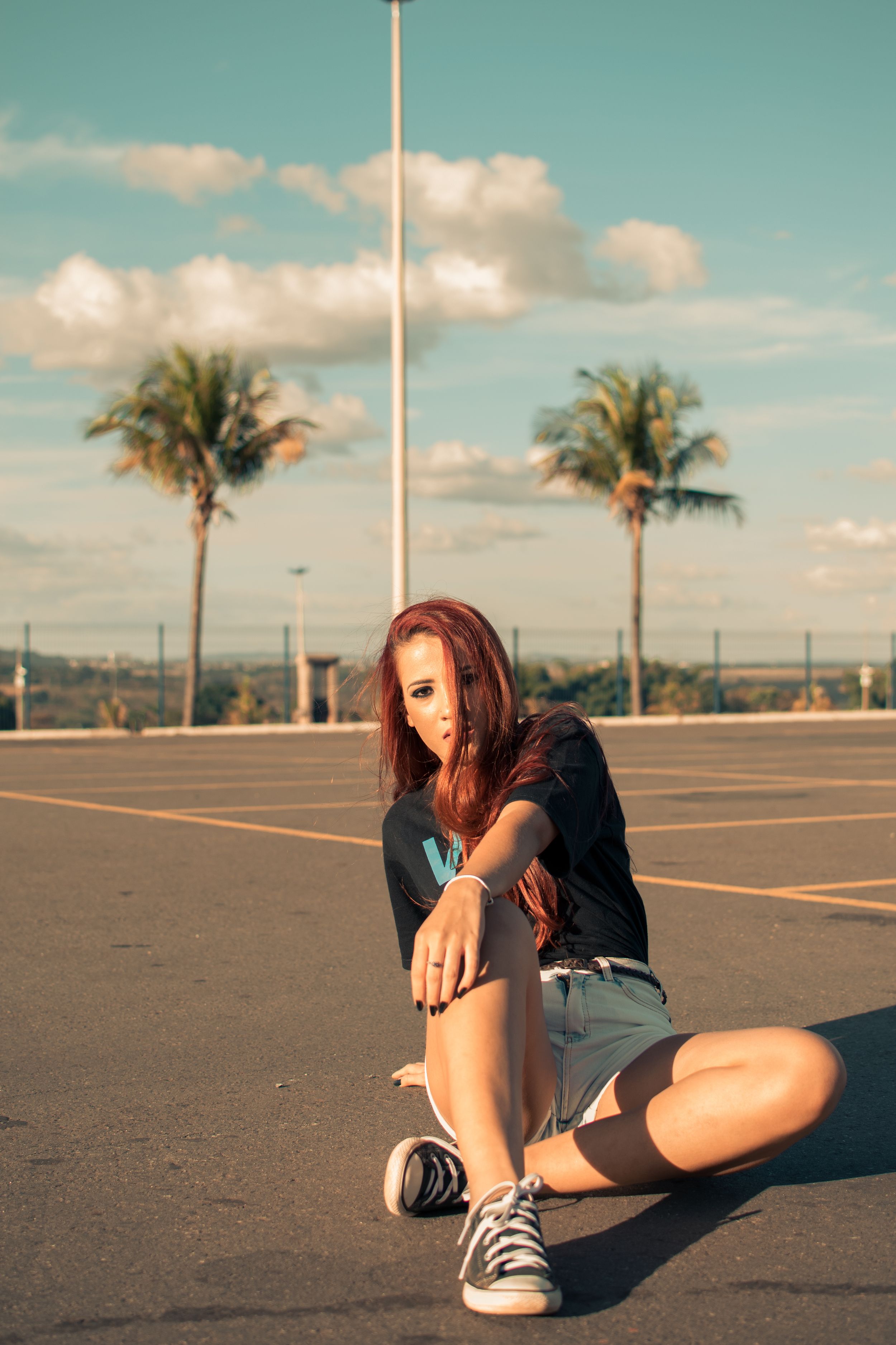 Woman in casual wear sitting in empty parking lot with palm trees and blue sky.