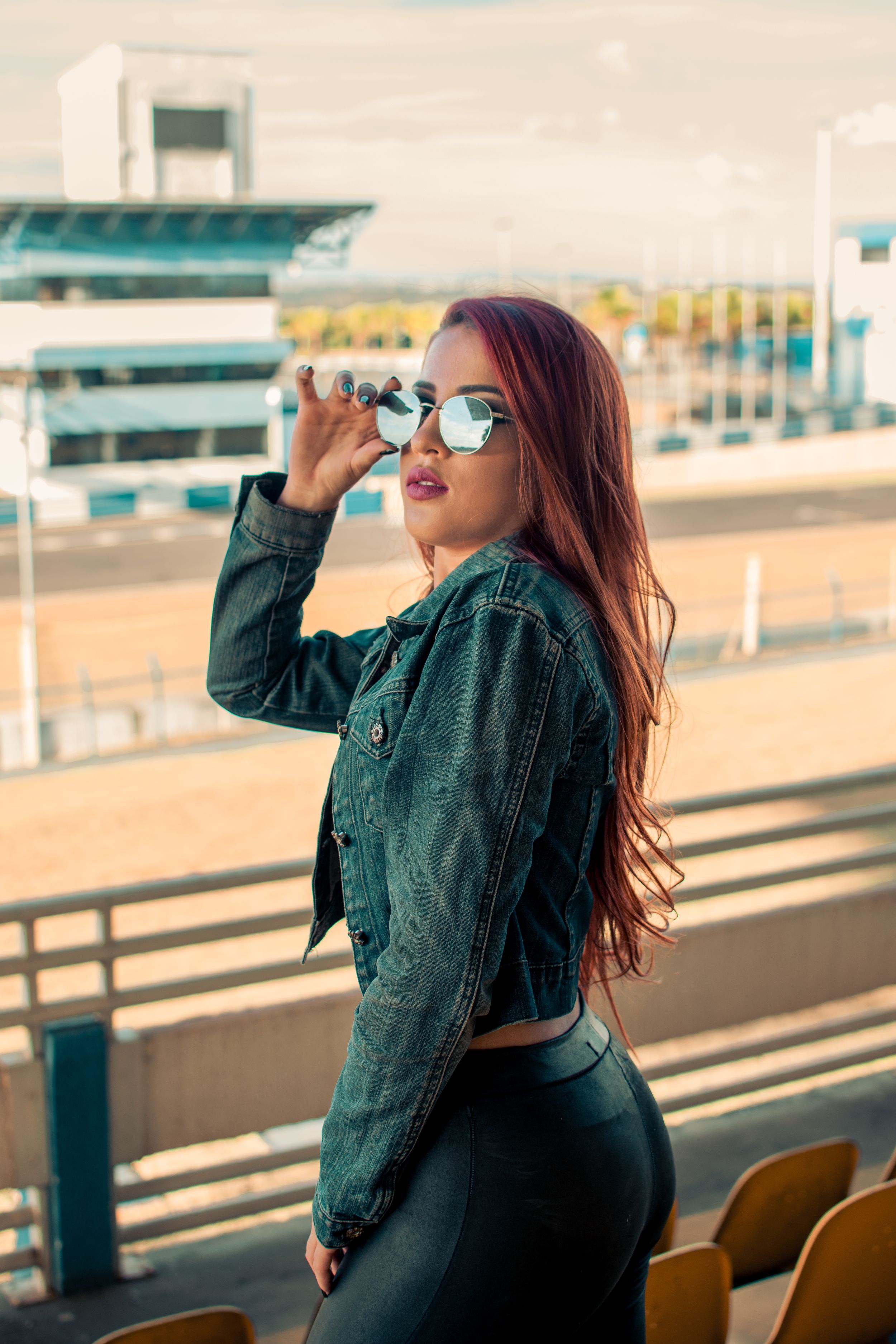 Side view of woman in denim jacket and sunglasses posing at stadium during golden hour.