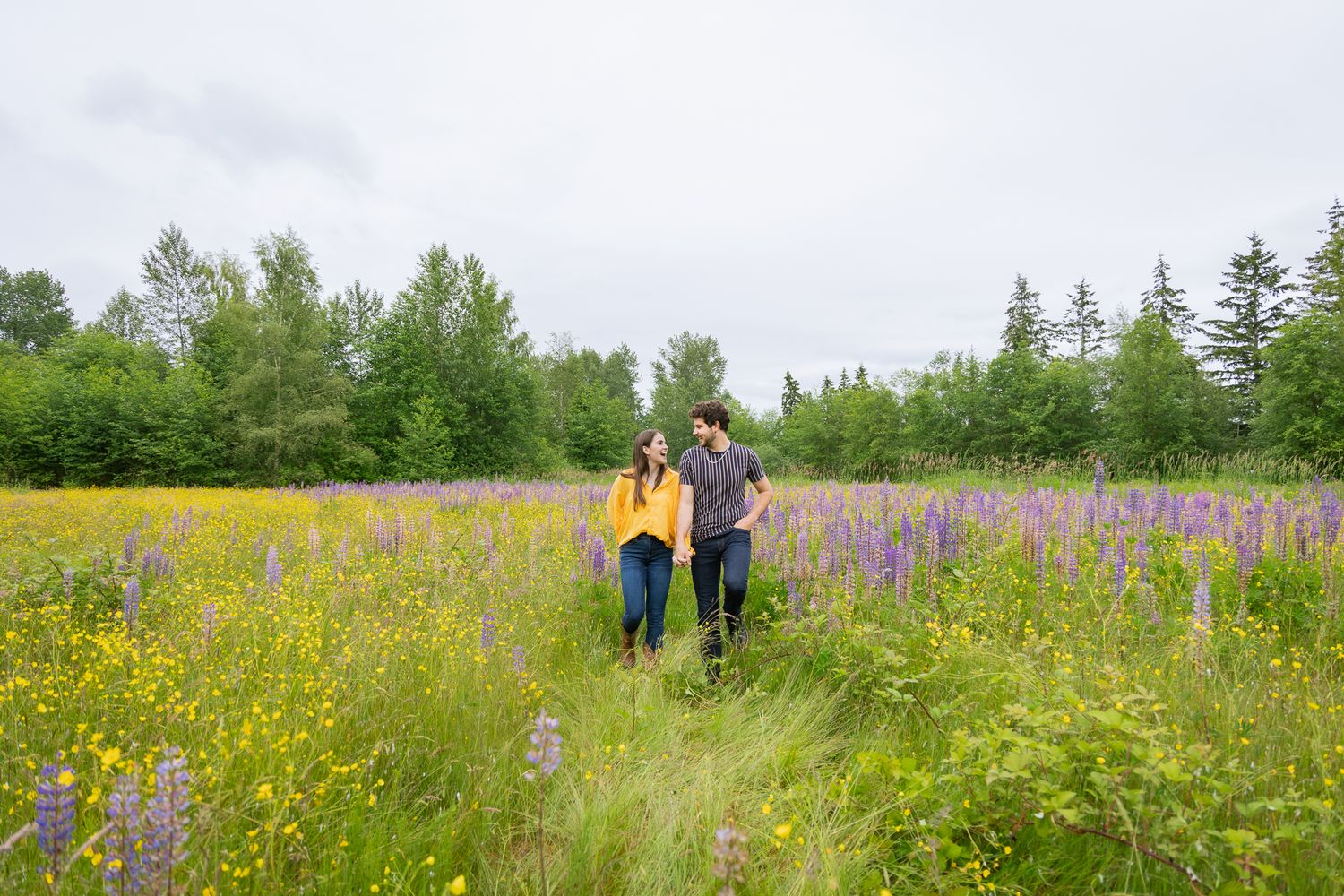 A couple walks towards camera, hand in hand, in a field of green with lots of purple flowers. They smile