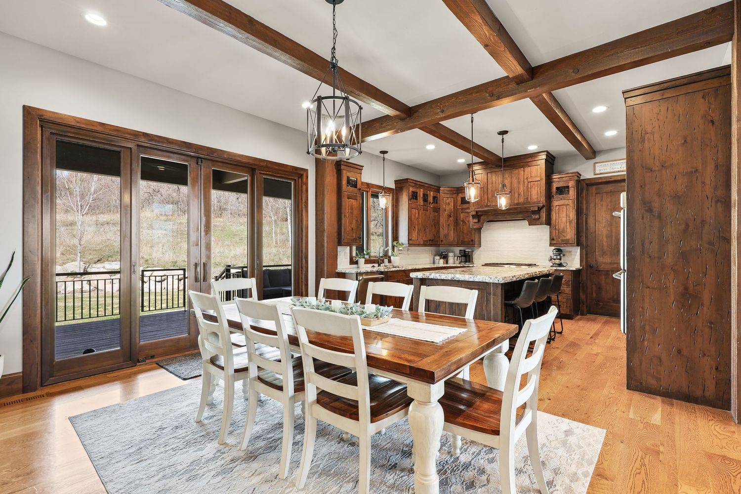 Elegant dining room with coffered ceiling, rustic wood table, white chairs, and open kitchen beyond. Captured by Slate Media (best in UT)