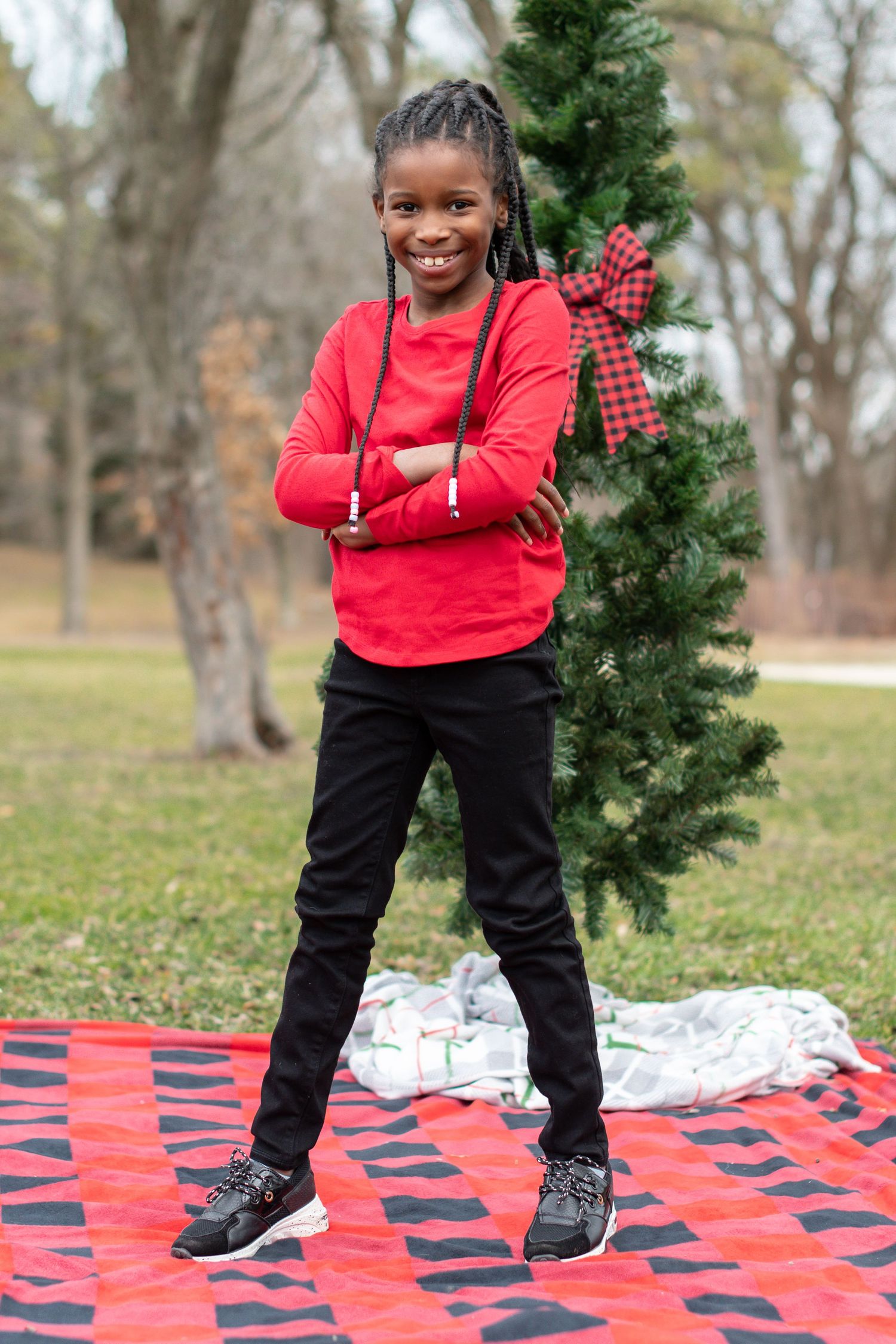 Child in a red sweater and black pants poses playfully on a red and black checkered blanket outdoors.