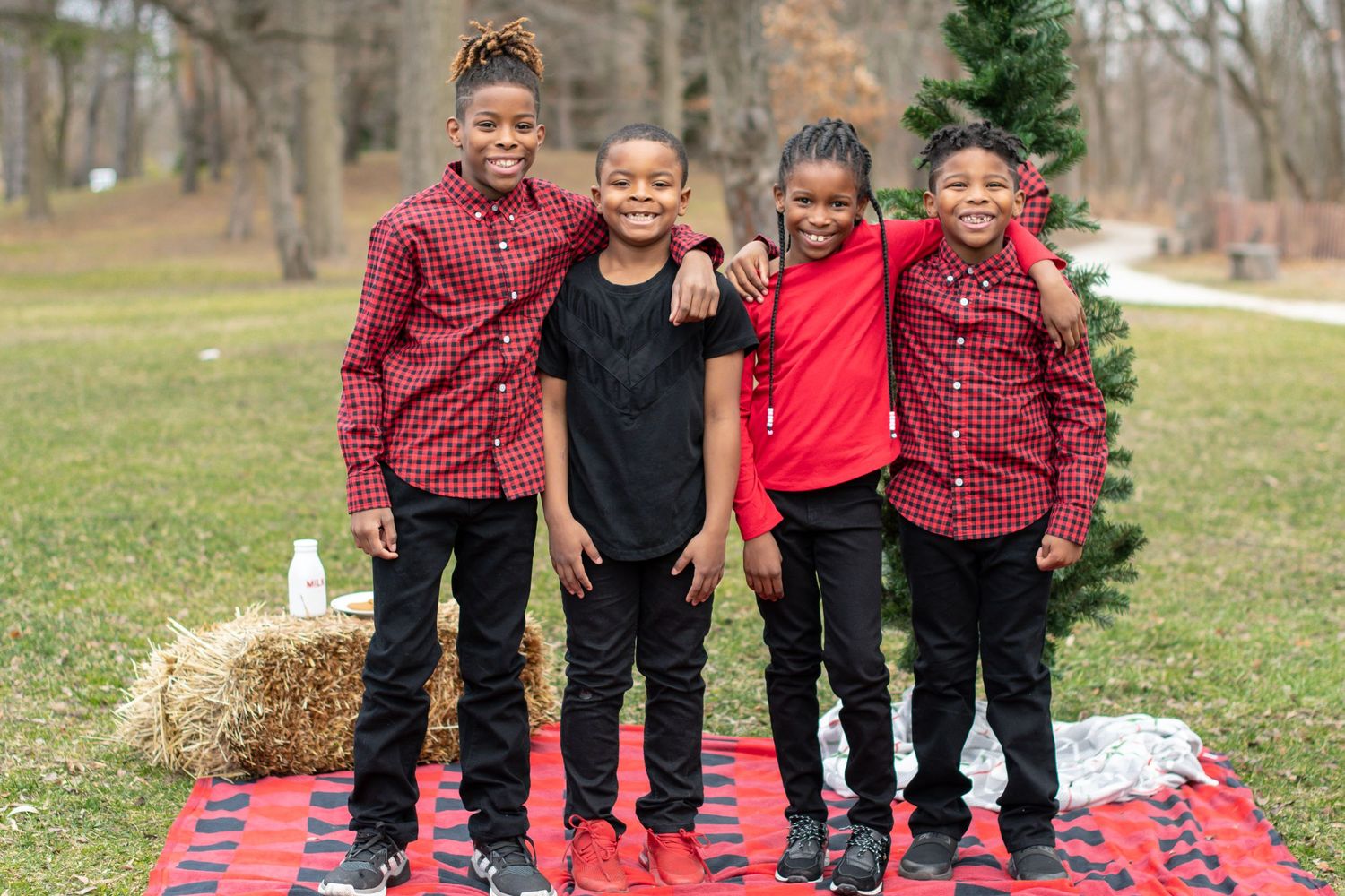 Group of small children stand together in coordinated red and black outfits near a Christmas tree.