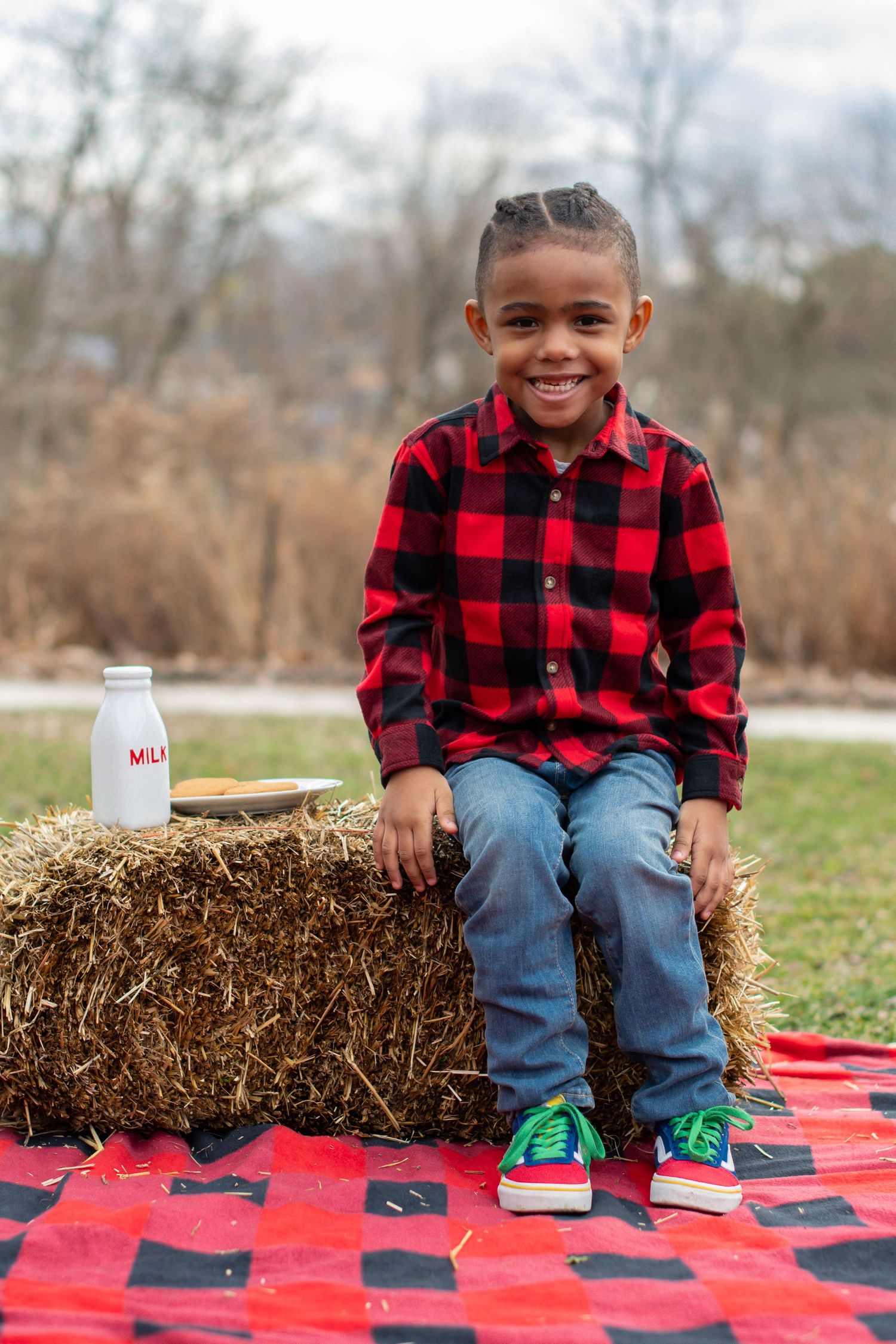 Child in buffalo plaid shirt sits on hay bale with milk bottle on red and black checkered blanket.