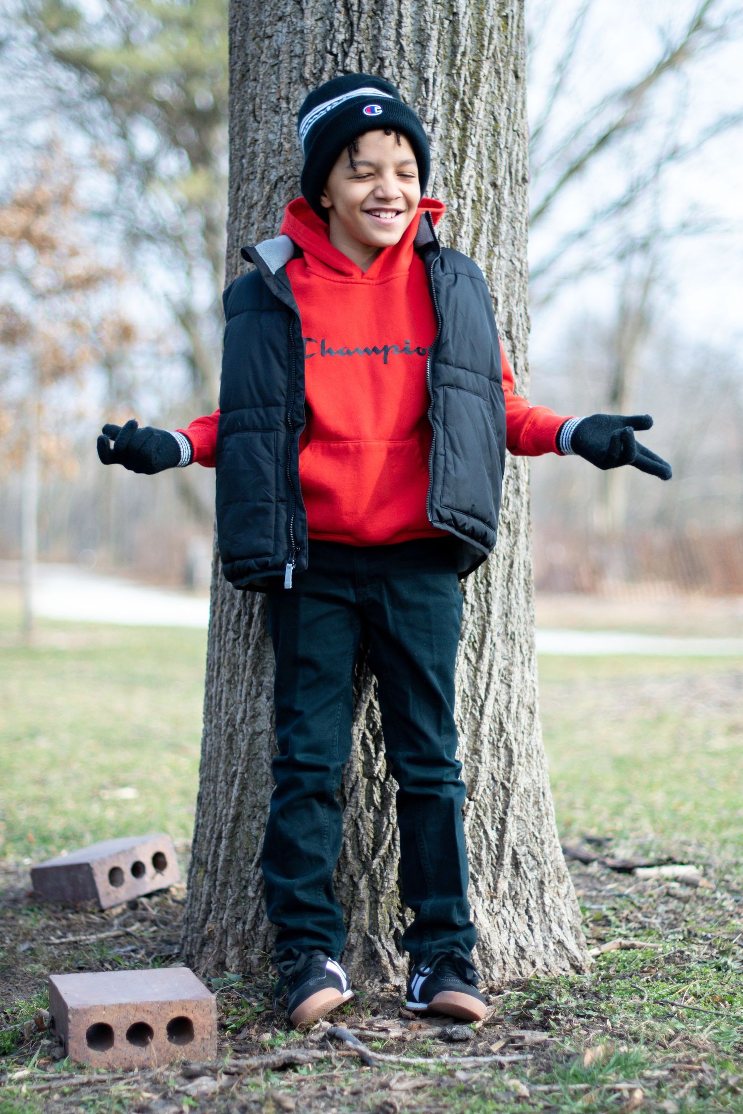 Young child playing outside and leaning against a tree trunk wearing red and black winter clothing.