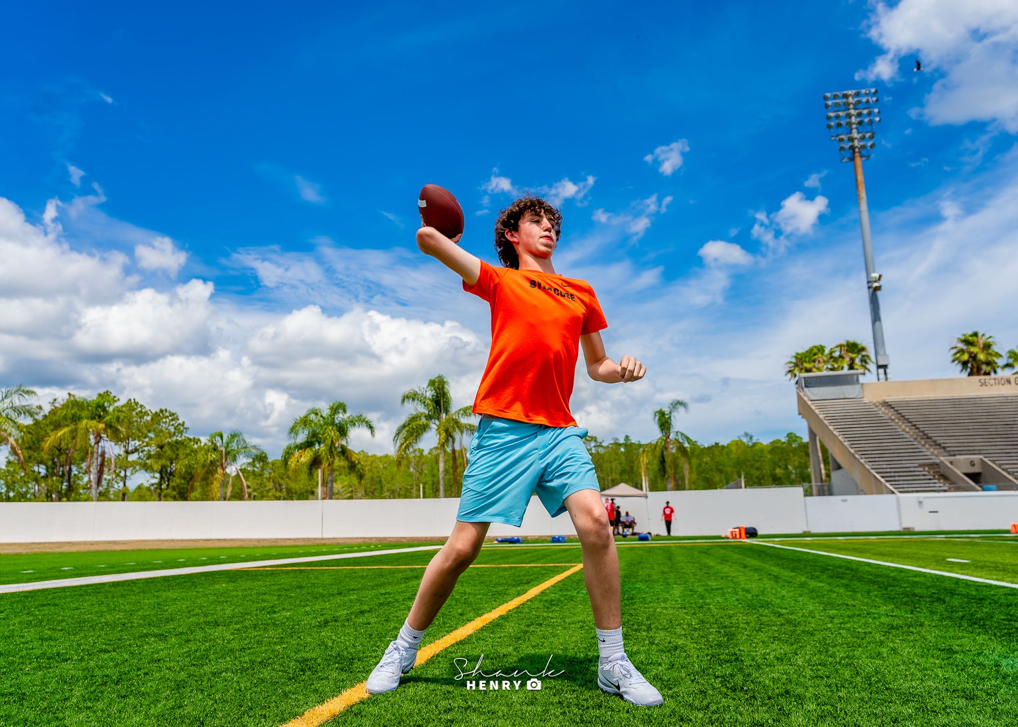 Young athlete practicing football throwing motion on sunny field with stadium in background.
