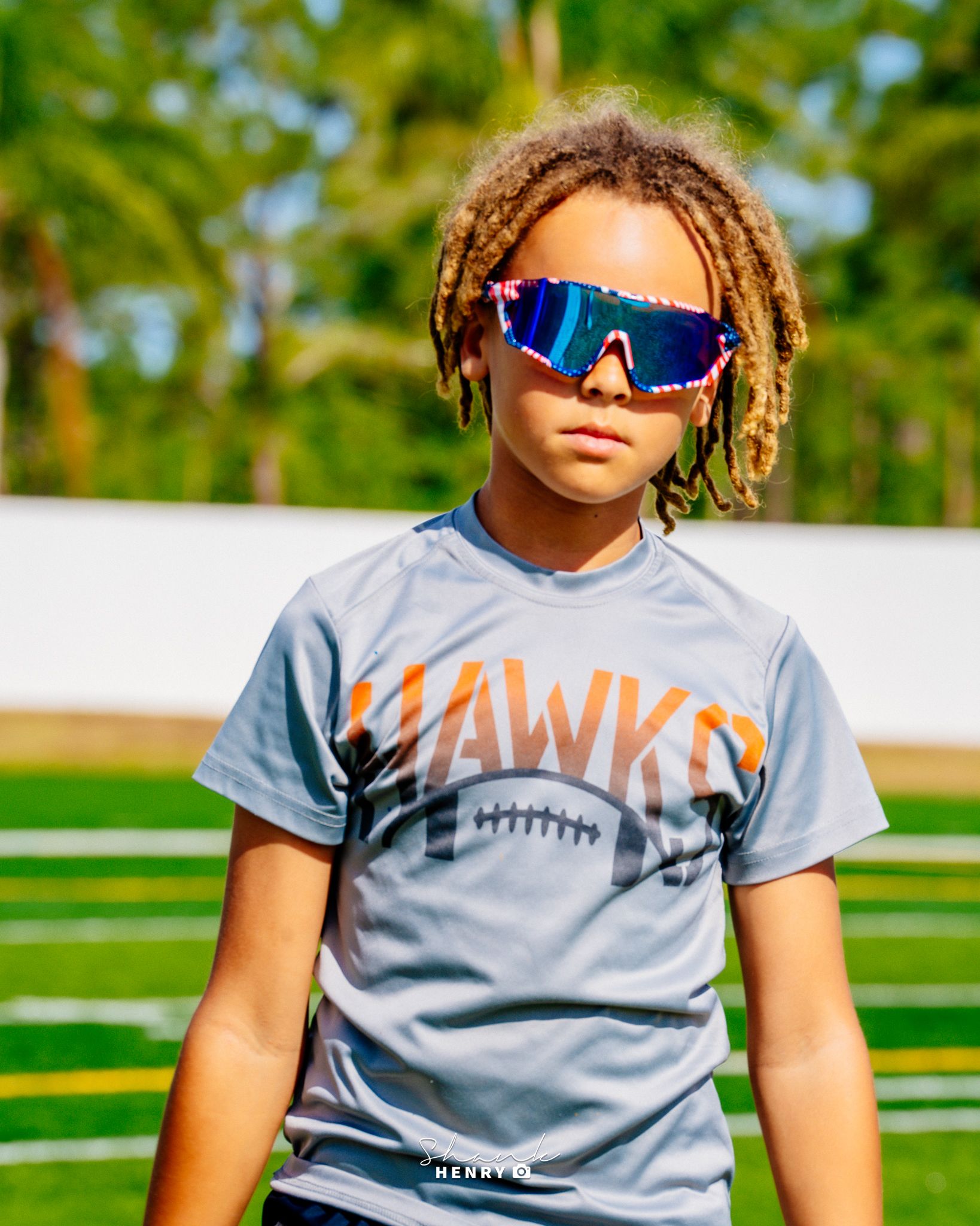 Young athlete in grey Hawks football shirt and sporty sunglasses on football field.