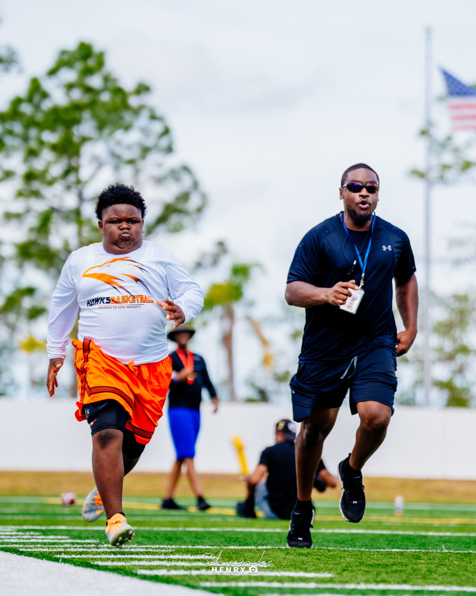 Two athletes sprint on a turf field with an American flag visible in the background.