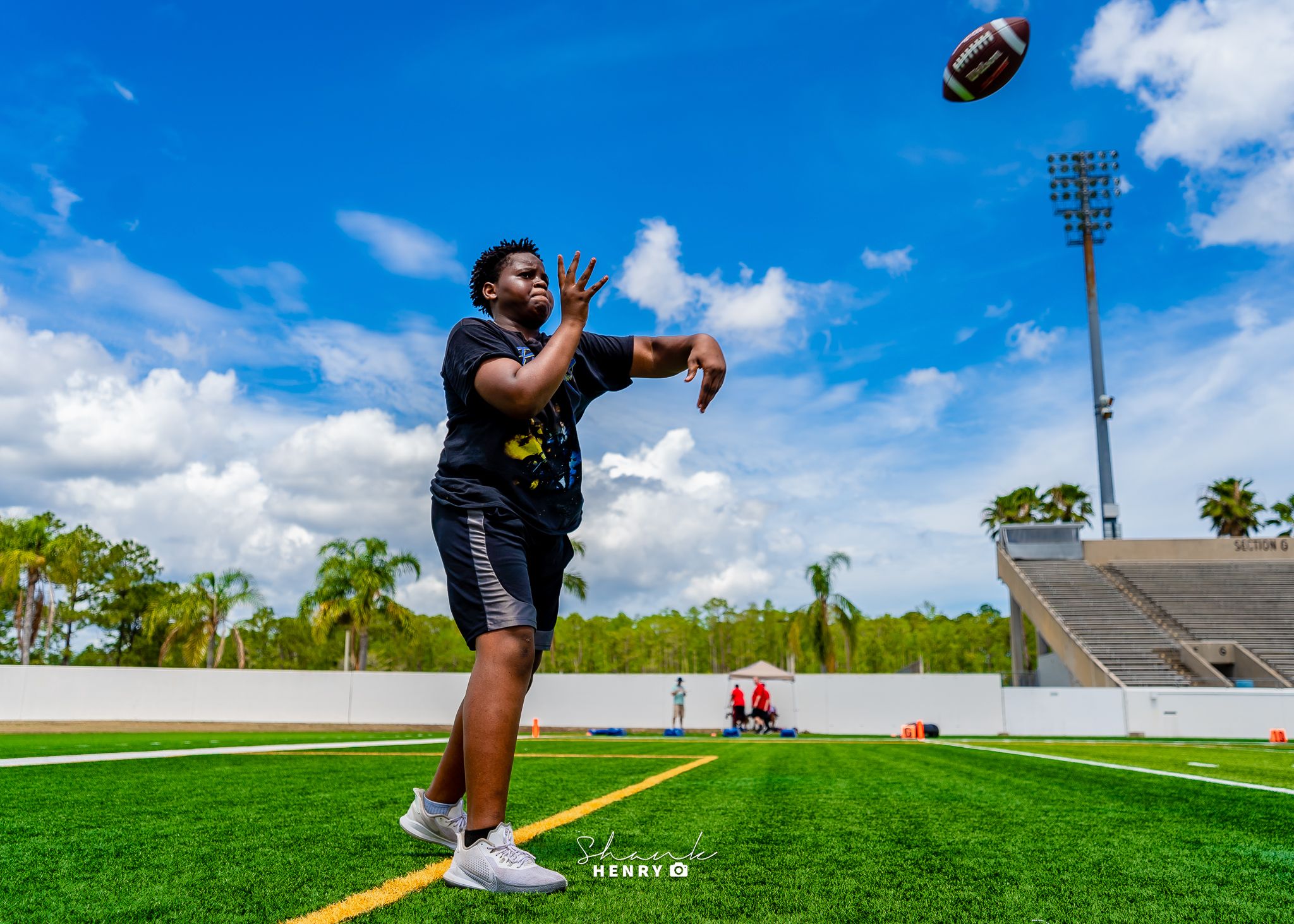 Football player practicing catching drills on a sunny day at an outdoor field with stadium lights in the background.