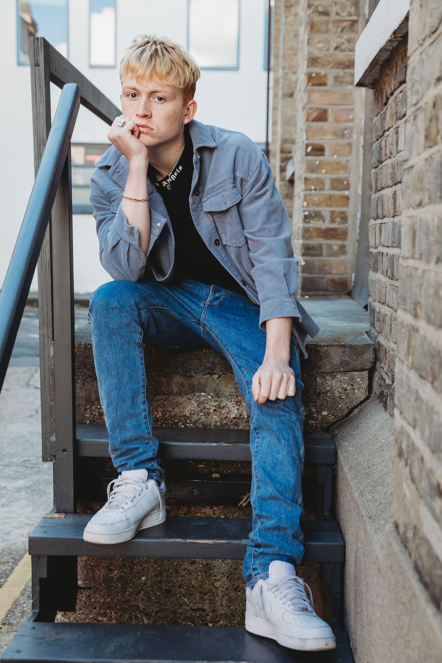 Person in denim jacket and jeans sits thoughtfully on outdoor metal stairs, in Stoke Newington urban location.