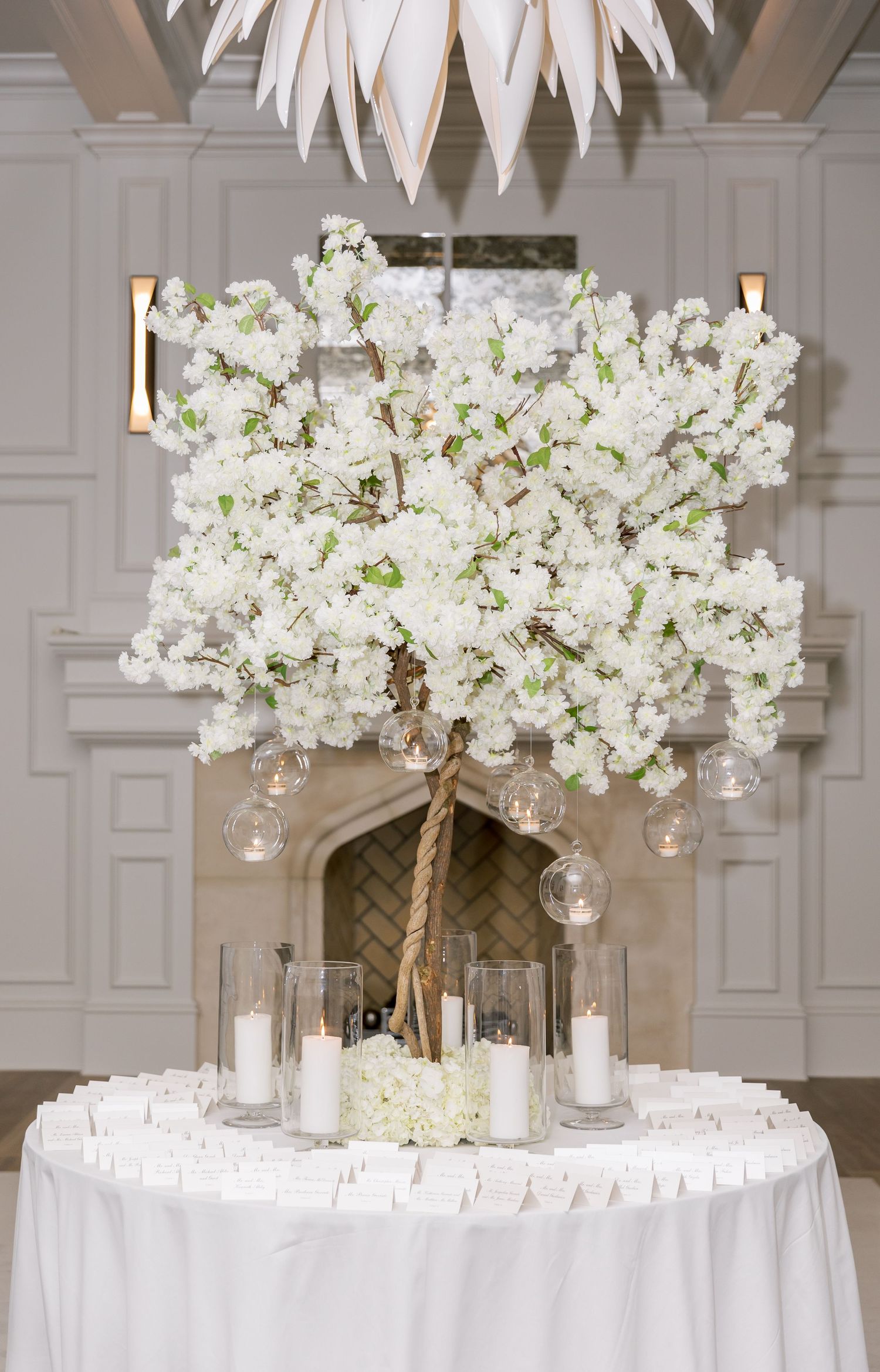 Stunning white floral centerpiece display with hanging glass orbs and white candles on round table.