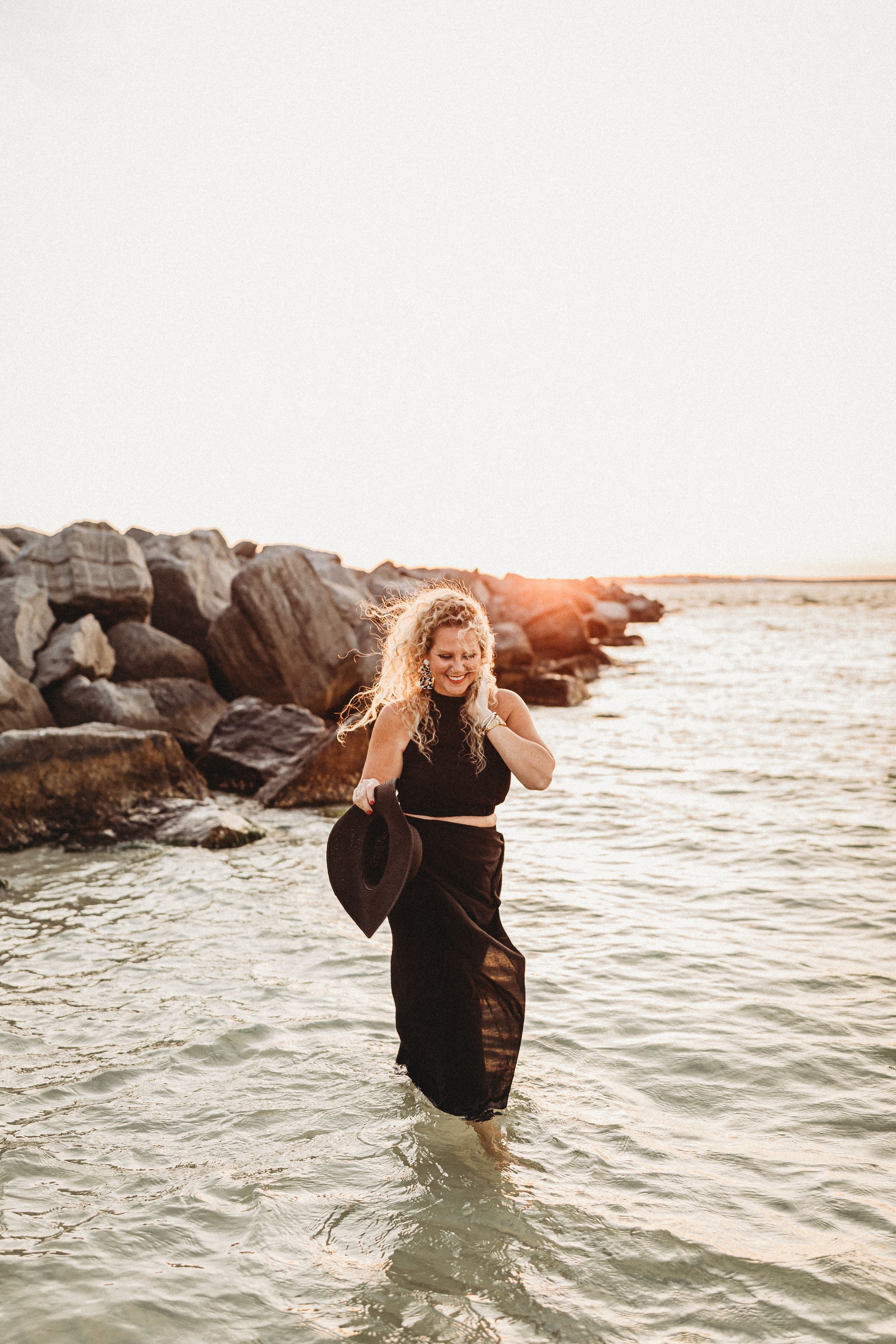 Woman in black dress wading through ocean water at sunset near rocky shore.
