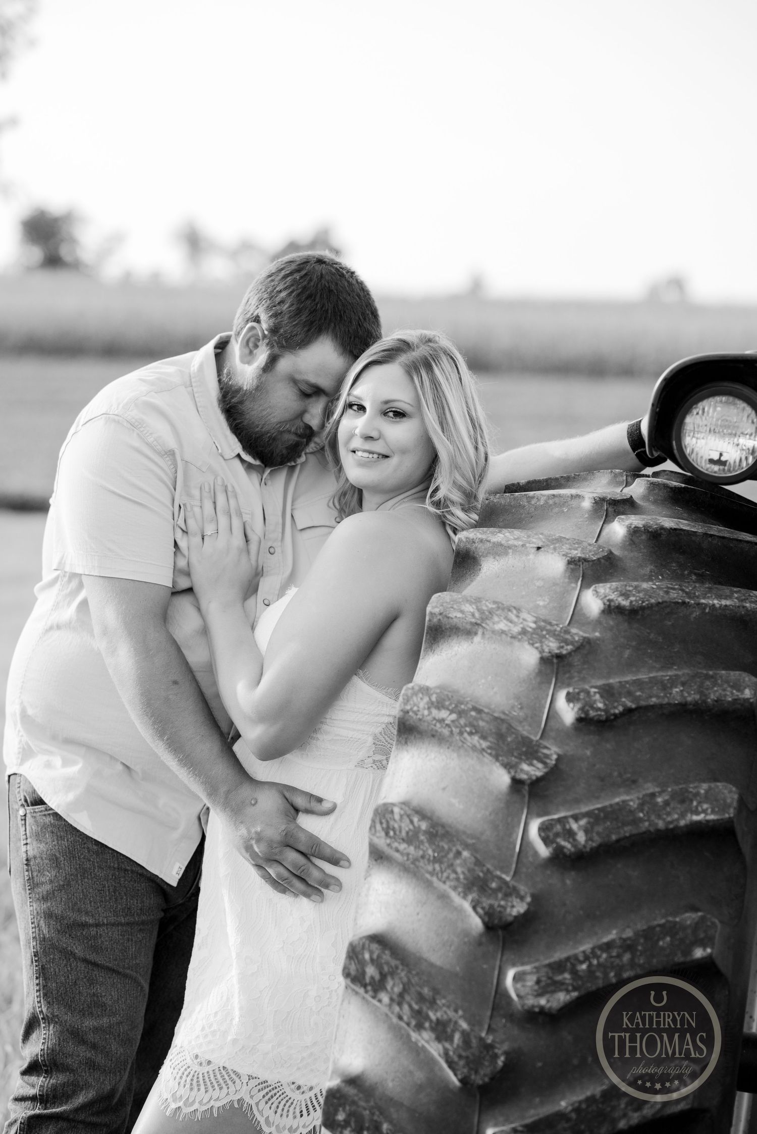 A couple poses romantically against the large tire of a vintage tractor in a black and white farm photo.