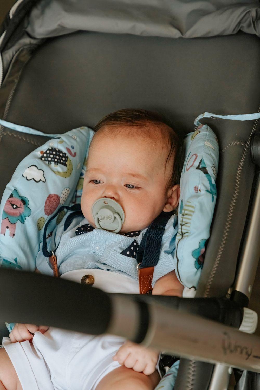 A baby rests comfortably in a stroller with light blue patterned padding and uses a pacifier.