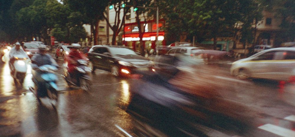 Blurred motion of traffic and scooters on a rainy city street at dusk.