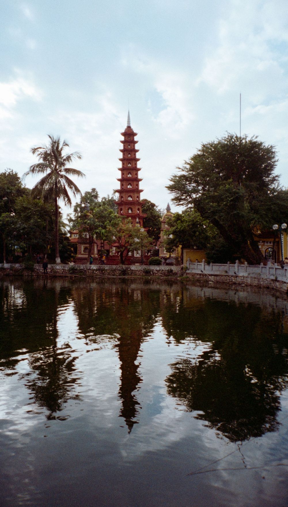Red pagoda temple reflecting in still water with palm trees and tropical foliage.
